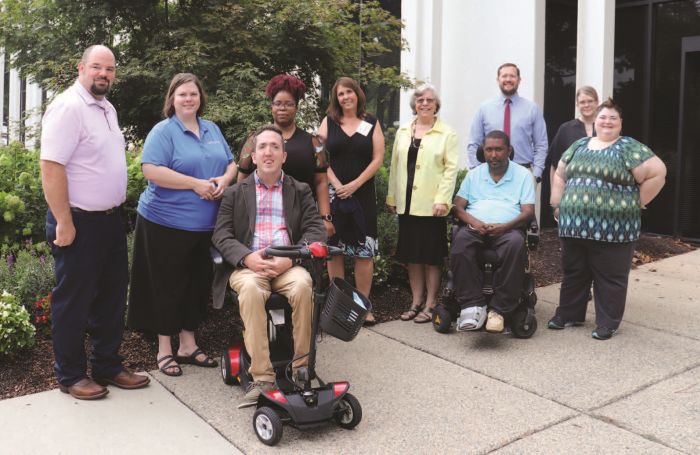 The SRC members gather on the sidewalk outside of the front entrance to the DARS Central Office. Green leafy trees and ground cover are in the background.