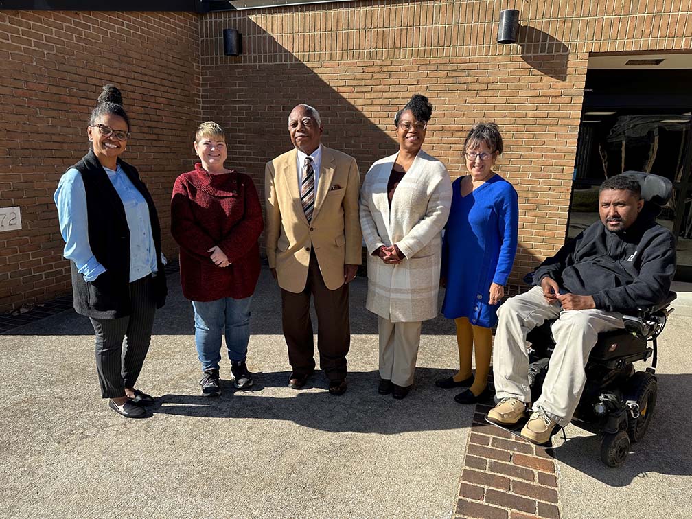 SRC members outside Wilson Workforce Rehabilitation Center in Fishersville, VA during a break at their November 2023 meeting. They stand in a corner niche of the red brick building in the bright direct sunlight, with some in long sleaves and others in coaats, on what looks like might be a cool day.